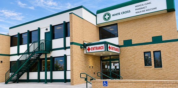 The White Cross Healthcare building at 647 Broadway in Winnipeg: a two-storey tan brick and white building with a green-trimmed White Cross sign (Medical Clinic, Pharmacy, Walk-ins Welcome) and a red-lettered Entrance canopy.